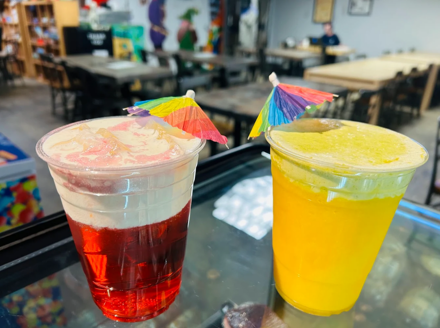 Two colorful drinks with umbrellas on a glass surface, with a game shop setting in the background.