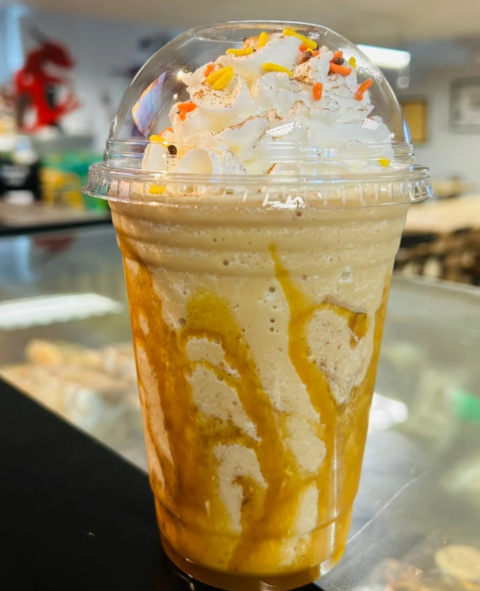 A frappuccino with whipped cream and sprinkles in a clear plastic cup on a reflective surface.